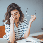 A woman suffering from the mental load of teaching. She is sitting at her desk, holding her glasses with one hand, and pinching the bridge of her nose with her other hand.