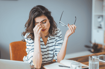 A woman suffering from the mental load of teaching. She is sitting at her desk, holding her glasses with one hand, and pinching the bridge of her nose with her other hand.